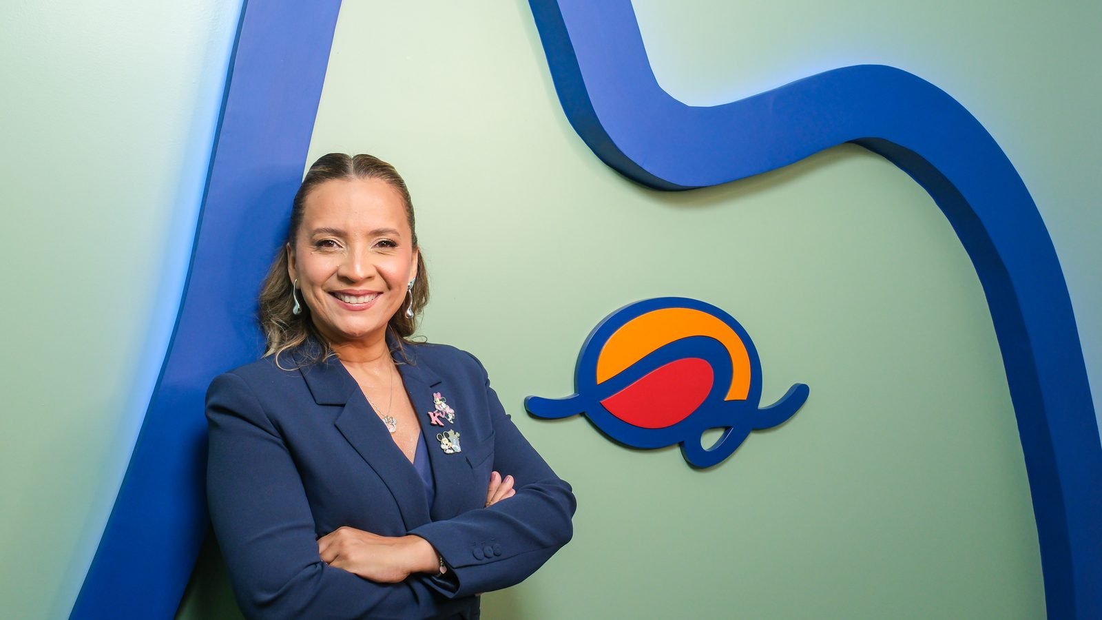 Kennya Martins de braços cruzados diante da parede verde, ao lado do símbolo do Instituto Mundo de Dentro
